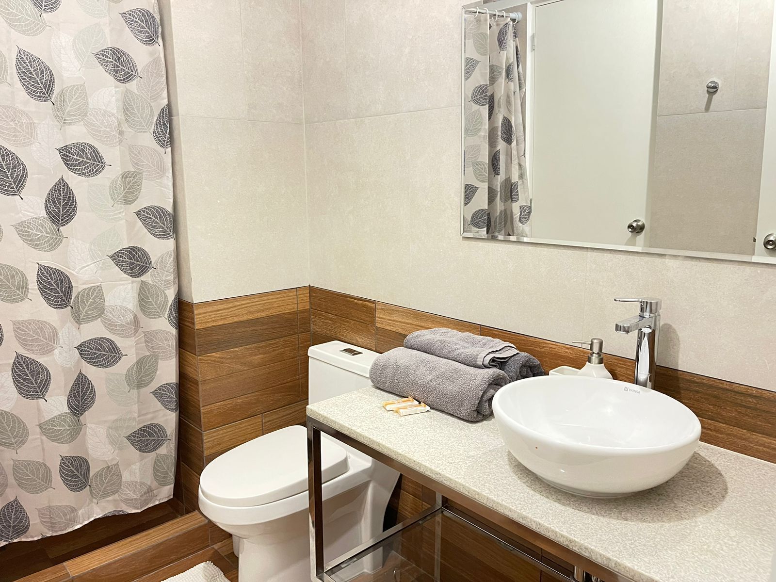 Un baño con inodoro, lavabo, espejo y cortina de ducha estampada con hojas.