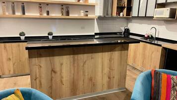 A modern kitchen with wooden cabinets, a black countertop, and a microwave. There are two blue chairs with colorful cushions.
