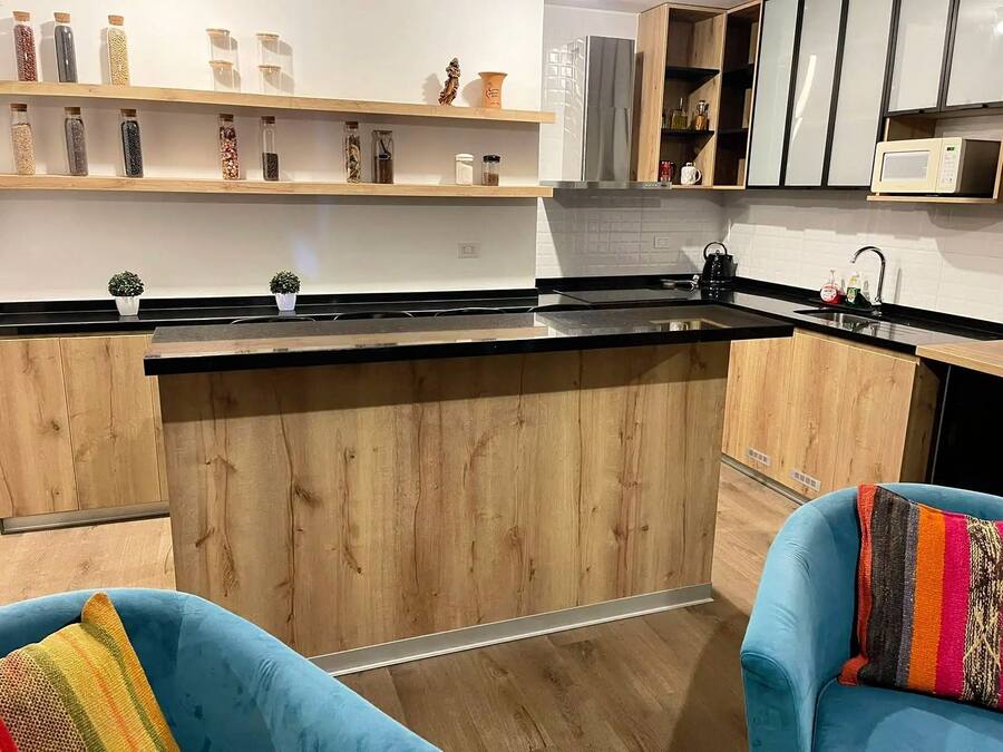 A modern kitchen with wooden cabinets, a black countertop, and a microwave. There are two blue chairs with colorful cushions.