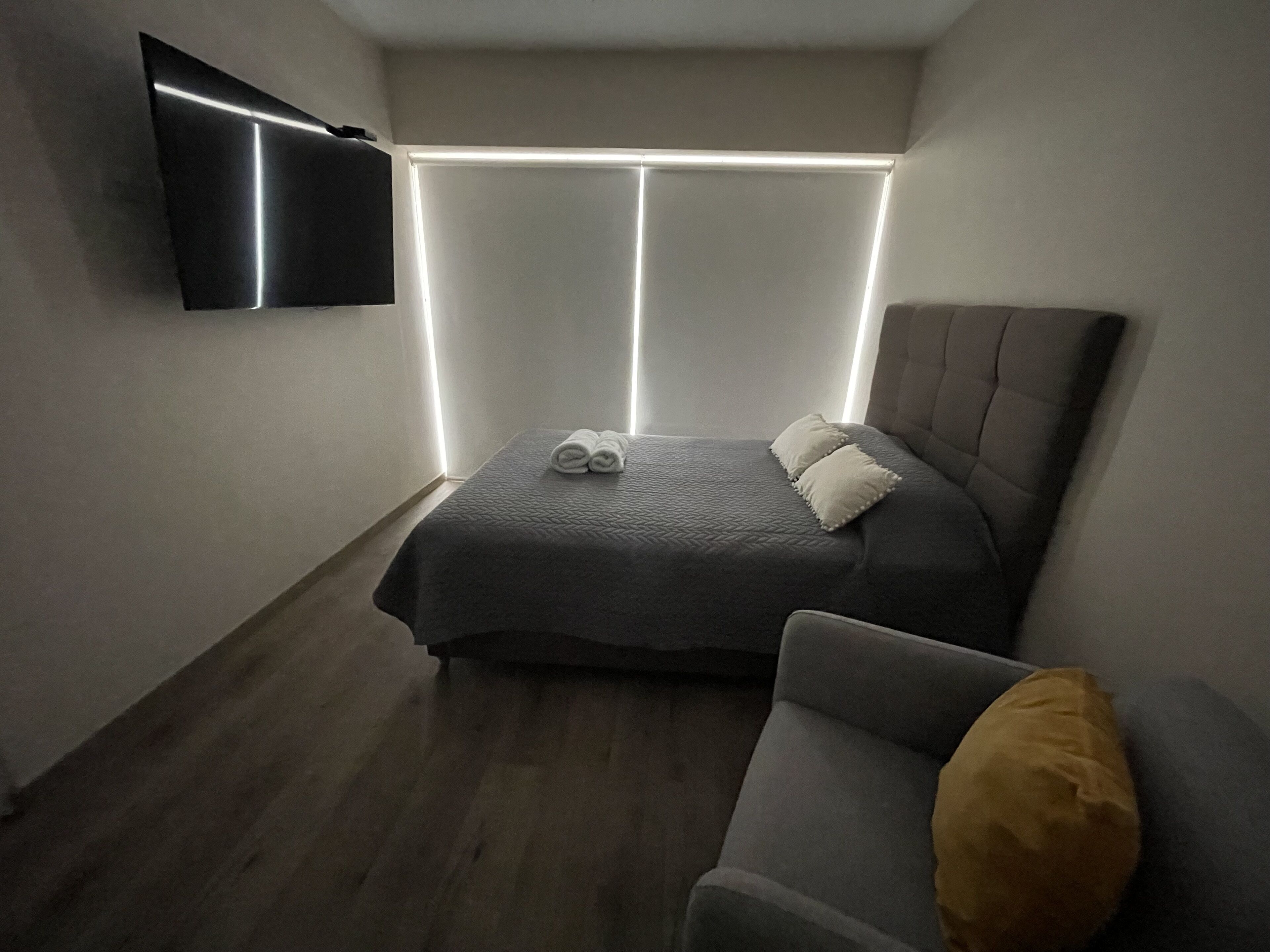 Una camera da letto con un letto, un comodino, una sedia, una TV a schermo piatto montata a parete e una finestra con persiane.