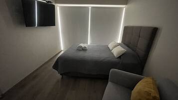 A bedroom with a bed, a bedside table, a chair, a flat-screen TV mounted on the wall, and a window with blinds.