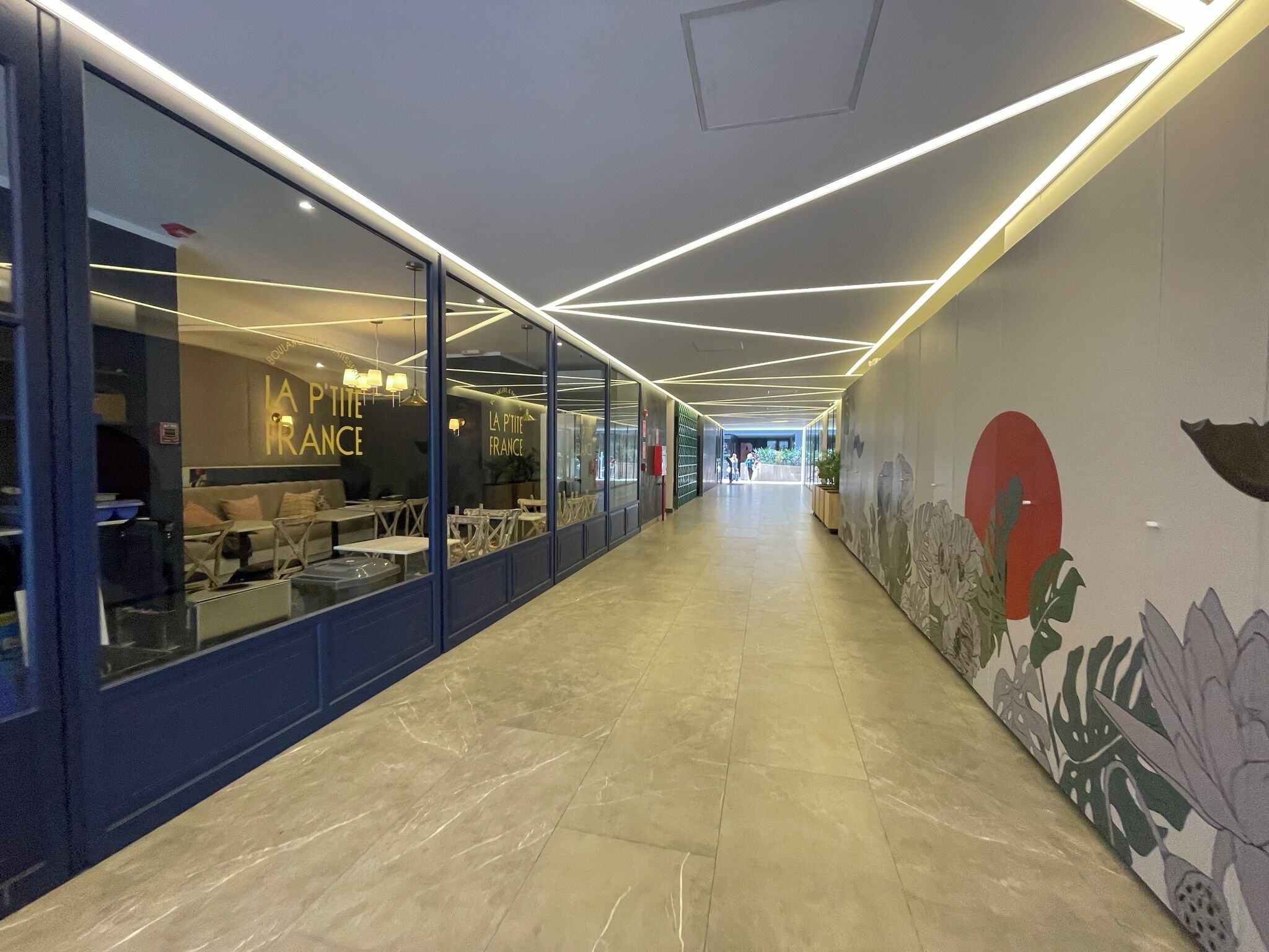 A corridor with glass walls, a tiled floor, and a ceiling with linear lighting.
