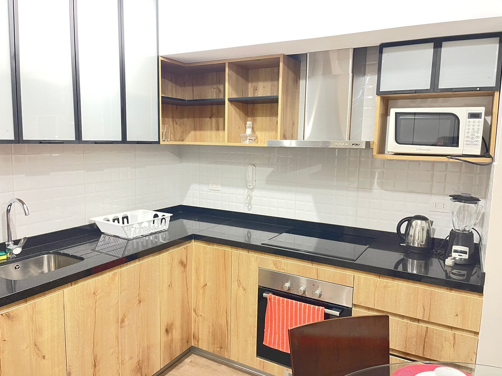 Una cocina moderna con armarios de madera, encimera negra, pileta de acero inoxidable, microondas, hervidor y licuadora.