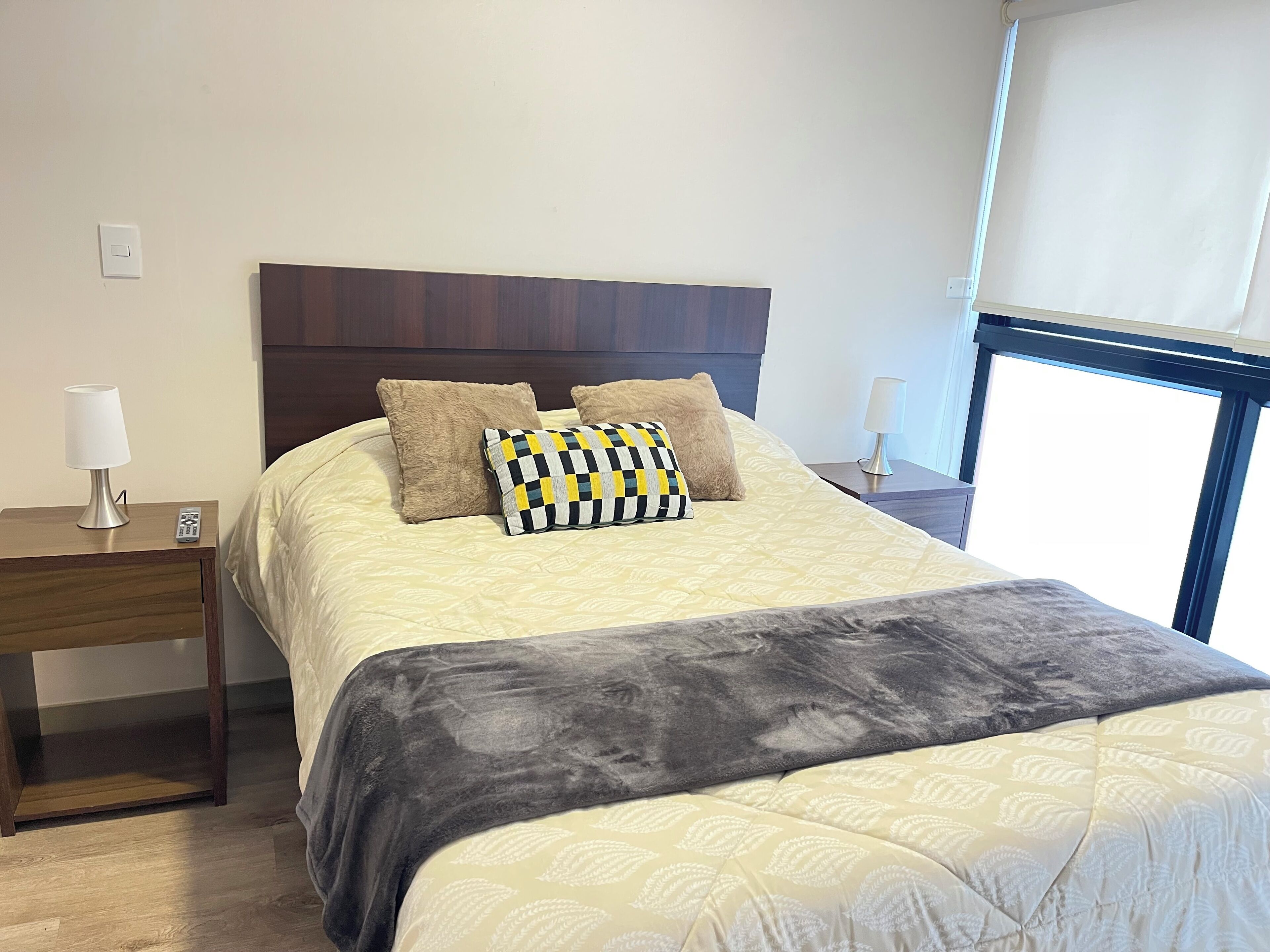 A neatly made bed with a patterned bedspread, two pillows, a dark throw blanket, and a nightstand with a lamp and remote control.