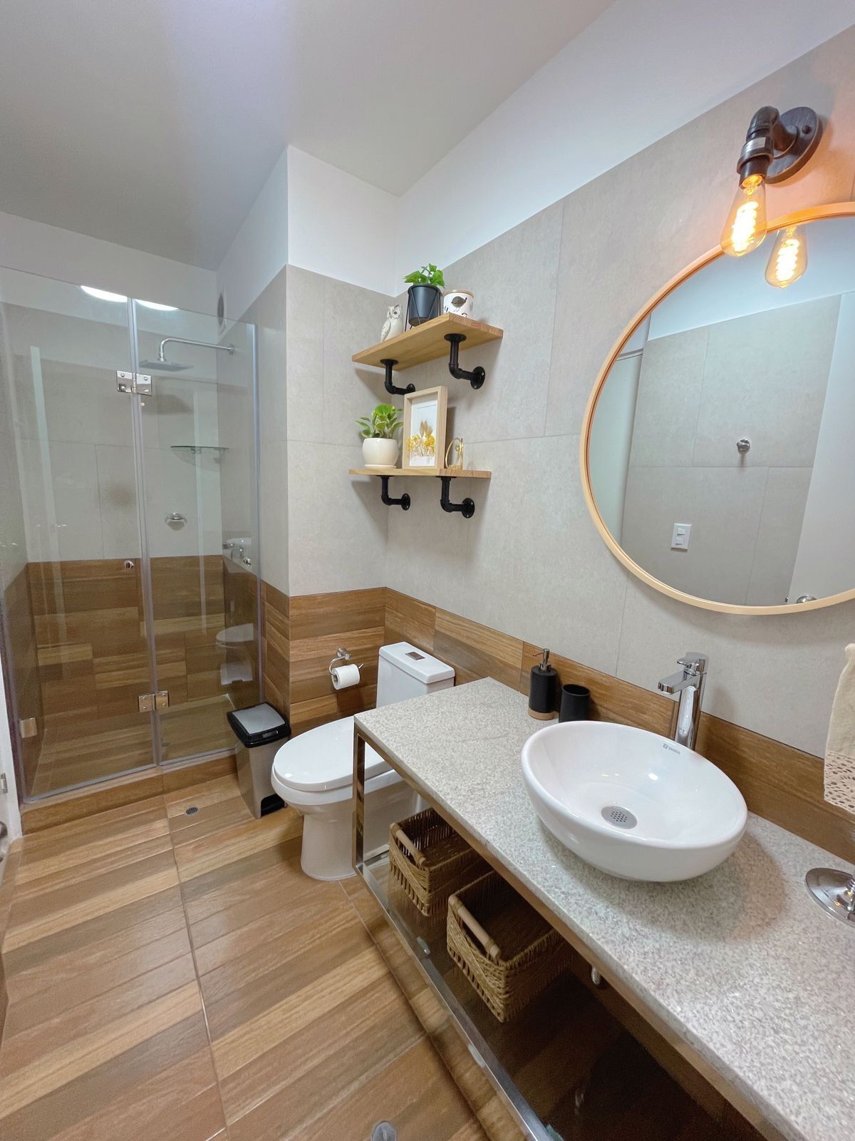 A modern bathroom with a glass-enclosed shower, a toilet, a sink, and a mirror. There are wooden shelves with decorative items and a round mirror on the wall.