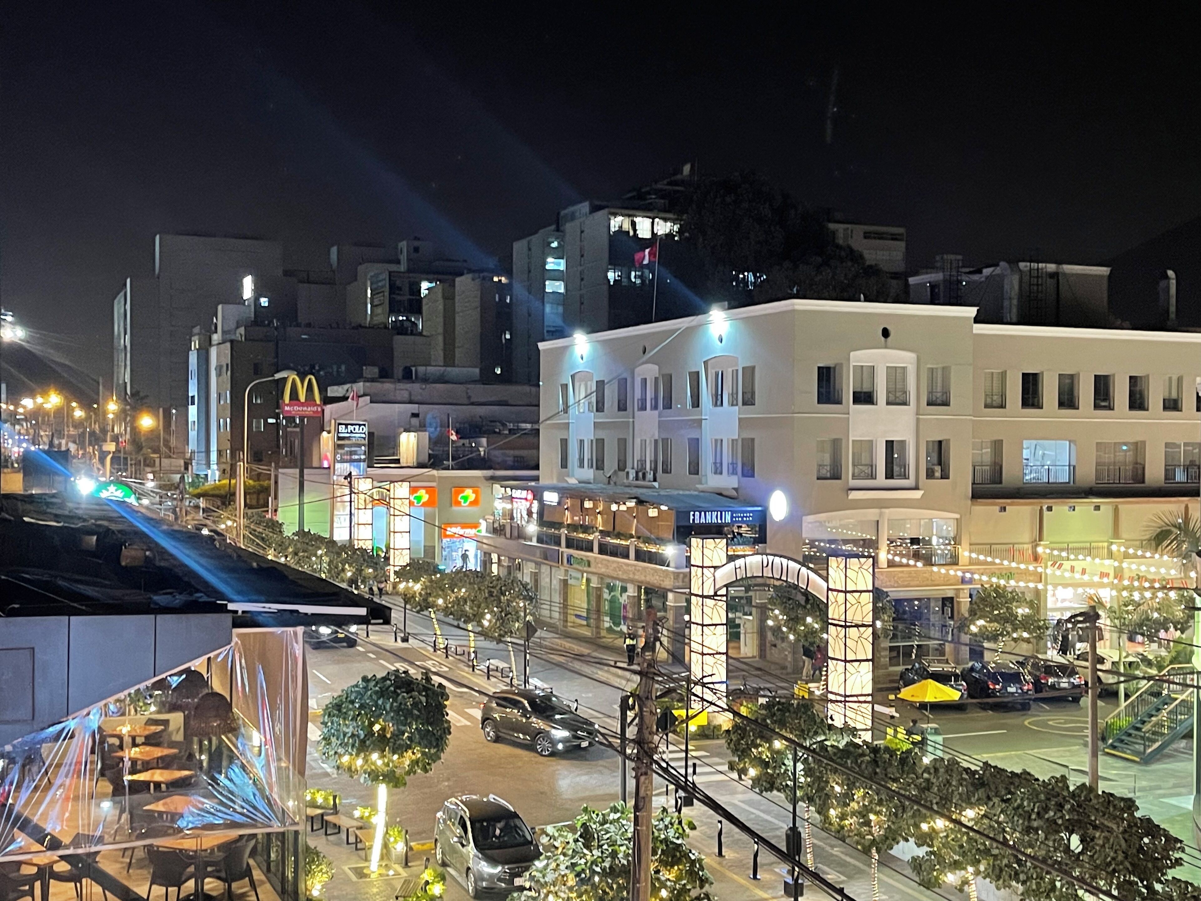 Un paisaje urbano nocturno con edificios iluminados, incluyendo un cartel llamativo de McDonald's, y una zona peatonal bien iluminada con árboles y mesas al aire libre.
