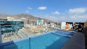 Une piscine à débordement située sur le toit, offrant une vue sur la ville, des bâtiments modernes et une chaîne de montagnes en arrière-plan.