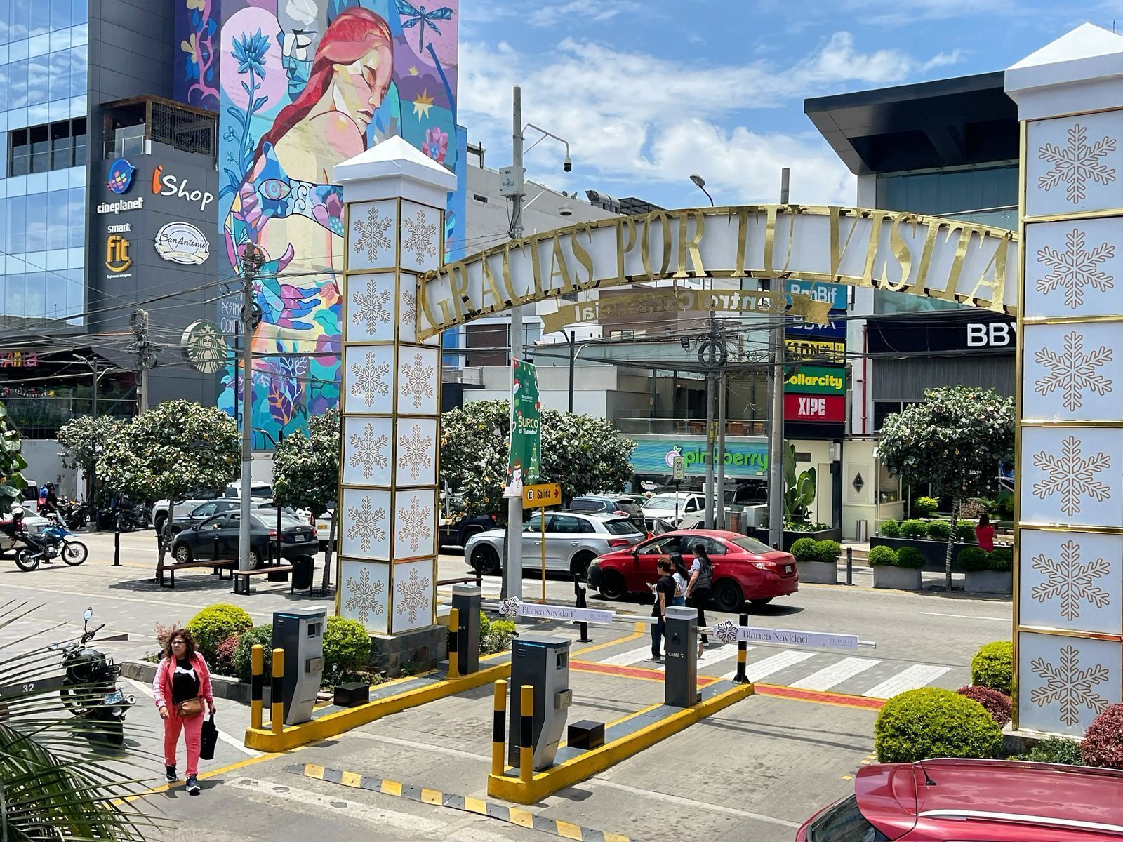 Una vivace strada cittadina con un grande murale su un edificio, una cabina di pedaggio e diversi negozi e veicoli.