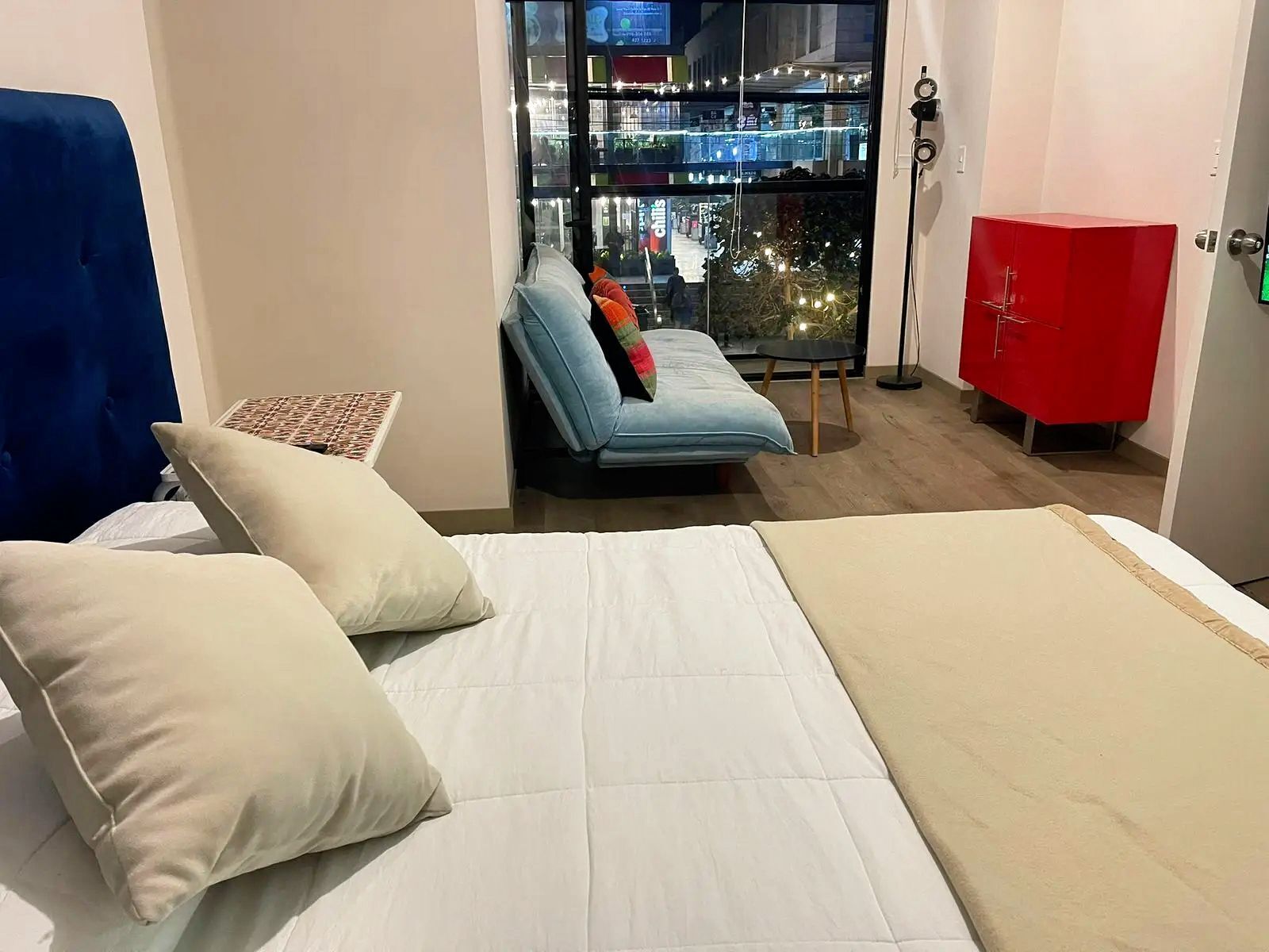 A hotel room with a bed, a blue chair, a red cabinet, and a view of a cityscape through a window.