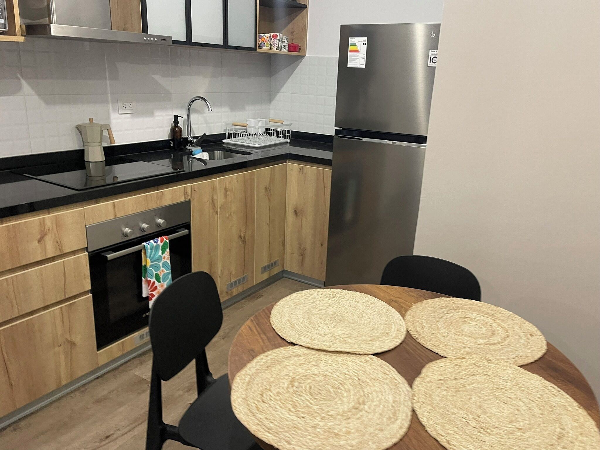 A modern kitchen with wooden cabinets, a stainless steel refrigerator, a built-in oven, and a round table with woven placemats.