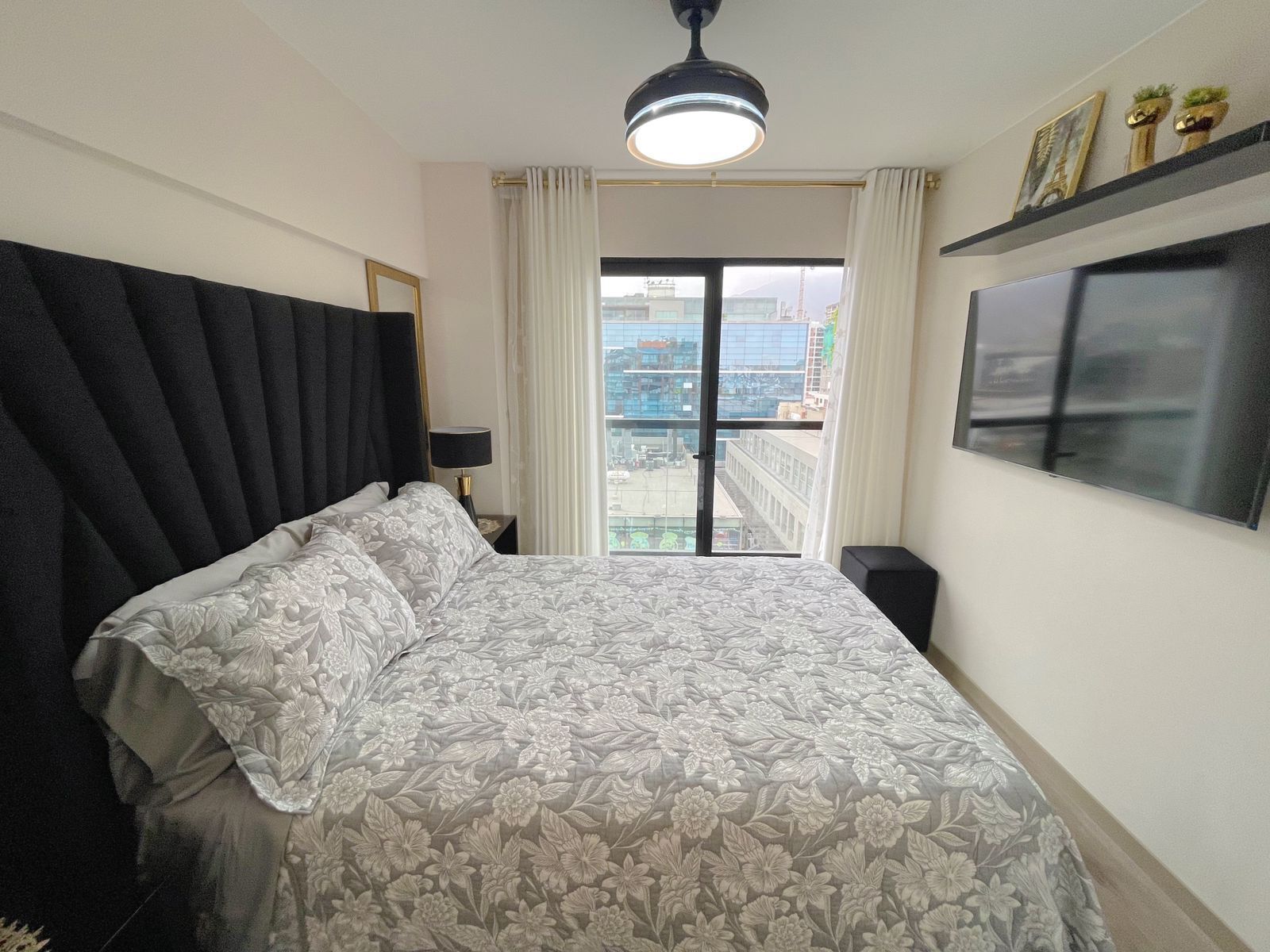 Ein Schlafzimmer mit einem großen Bett, einem an der Wand befestigten Fernseher, einem Balkon mit Vorhängen und Blick auf die Stadt.