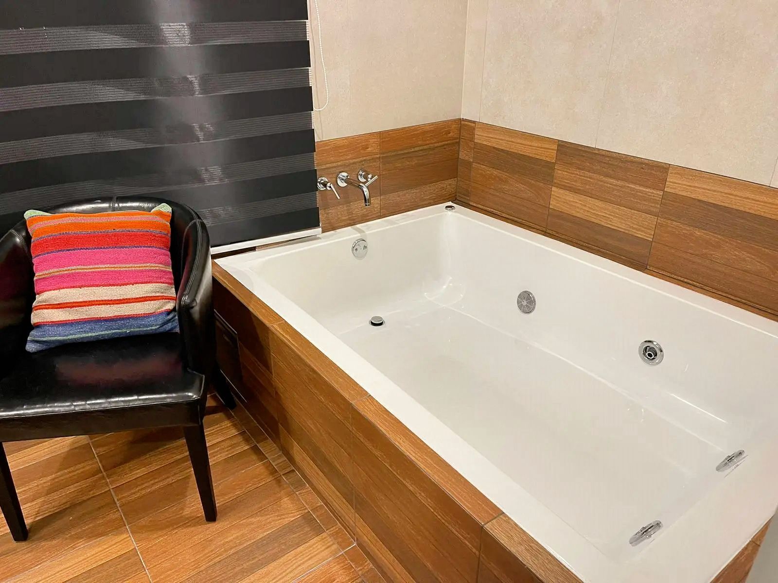 A modern bathroom with a wooden floor, a black chair, a colorful striped towel, and a white bathtub.