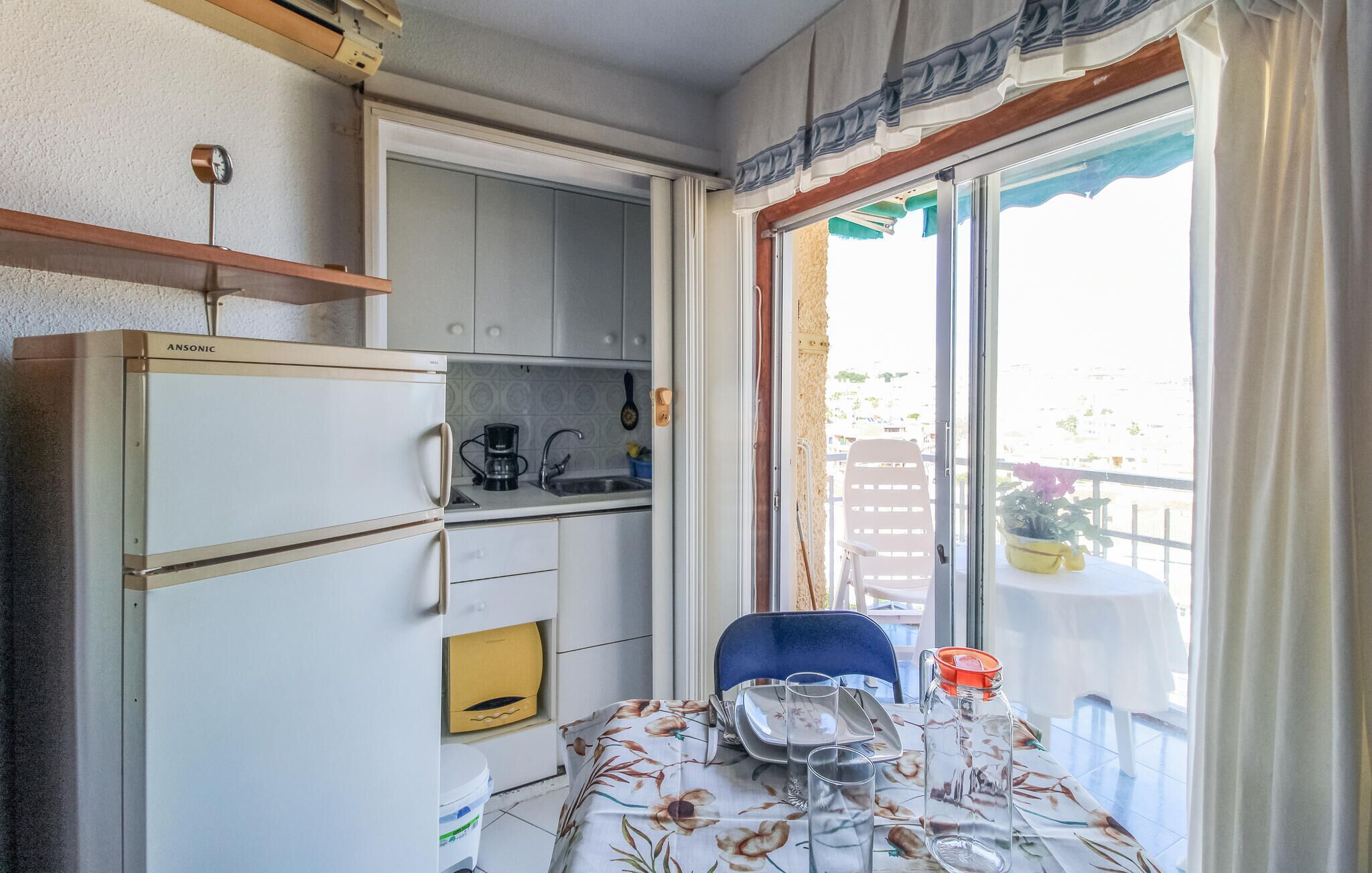 A compact kitchen with a coffee maker, sink, and storage cabinets. A dining table with a floral tablecloth and a chair is set up near a sliding glass door leading to an outdoor area.