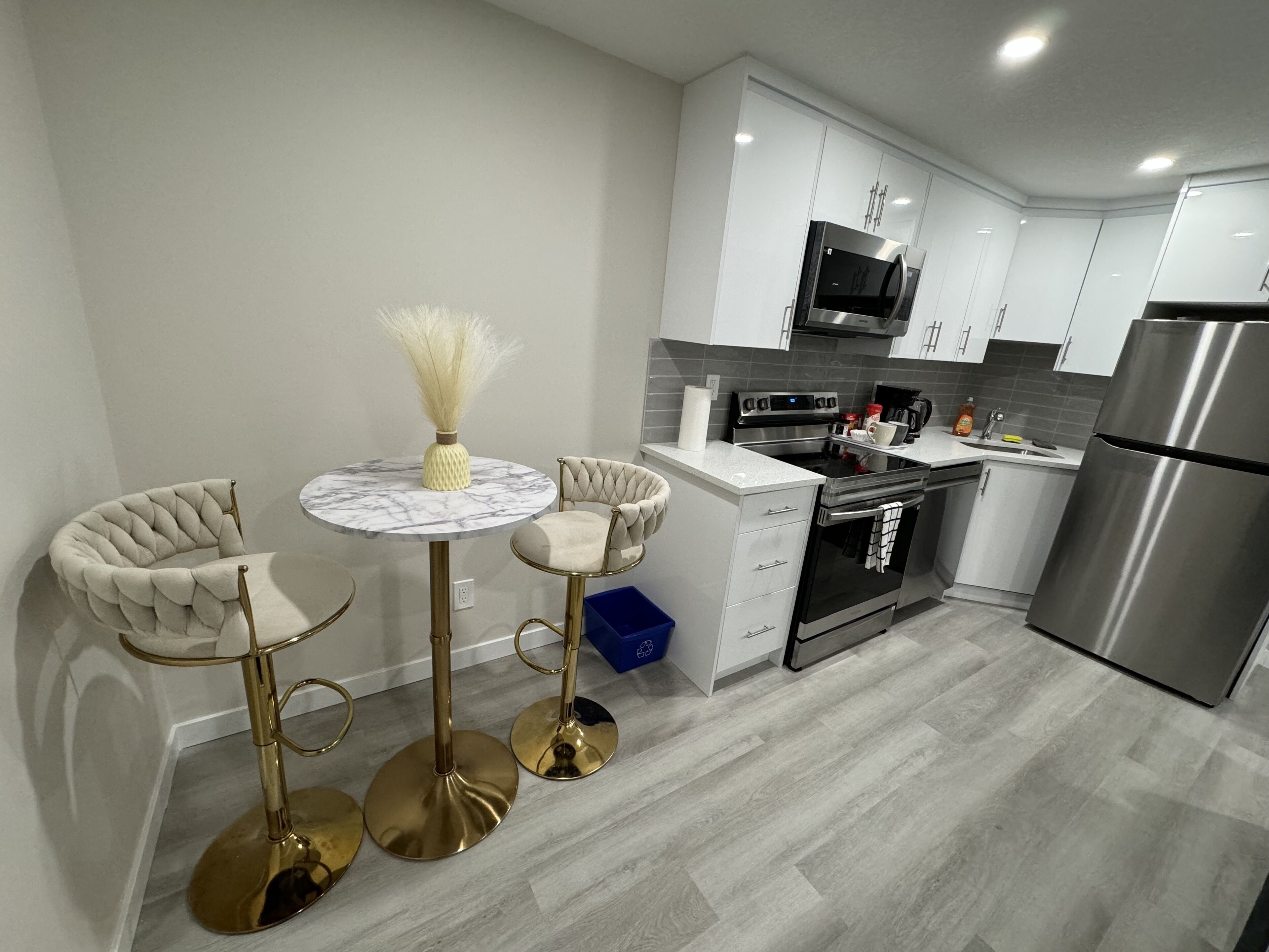 A modern kitchen with white cabinets, stainless steel appliances, and a marble-top dining table with two chairs.