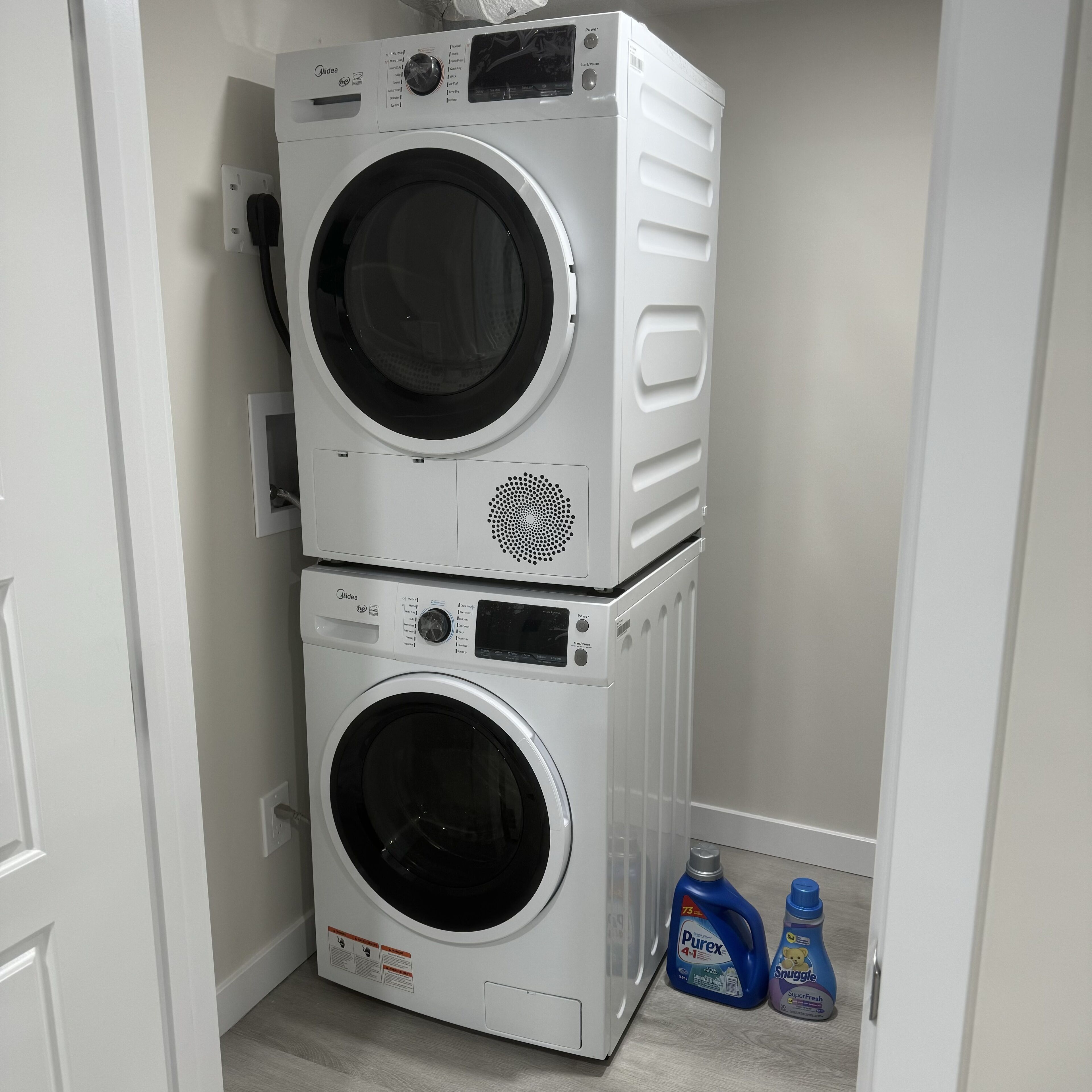 Two stacked front-loading washing machines in a laundry room with a door on the left and two detergent bottles on the floor.