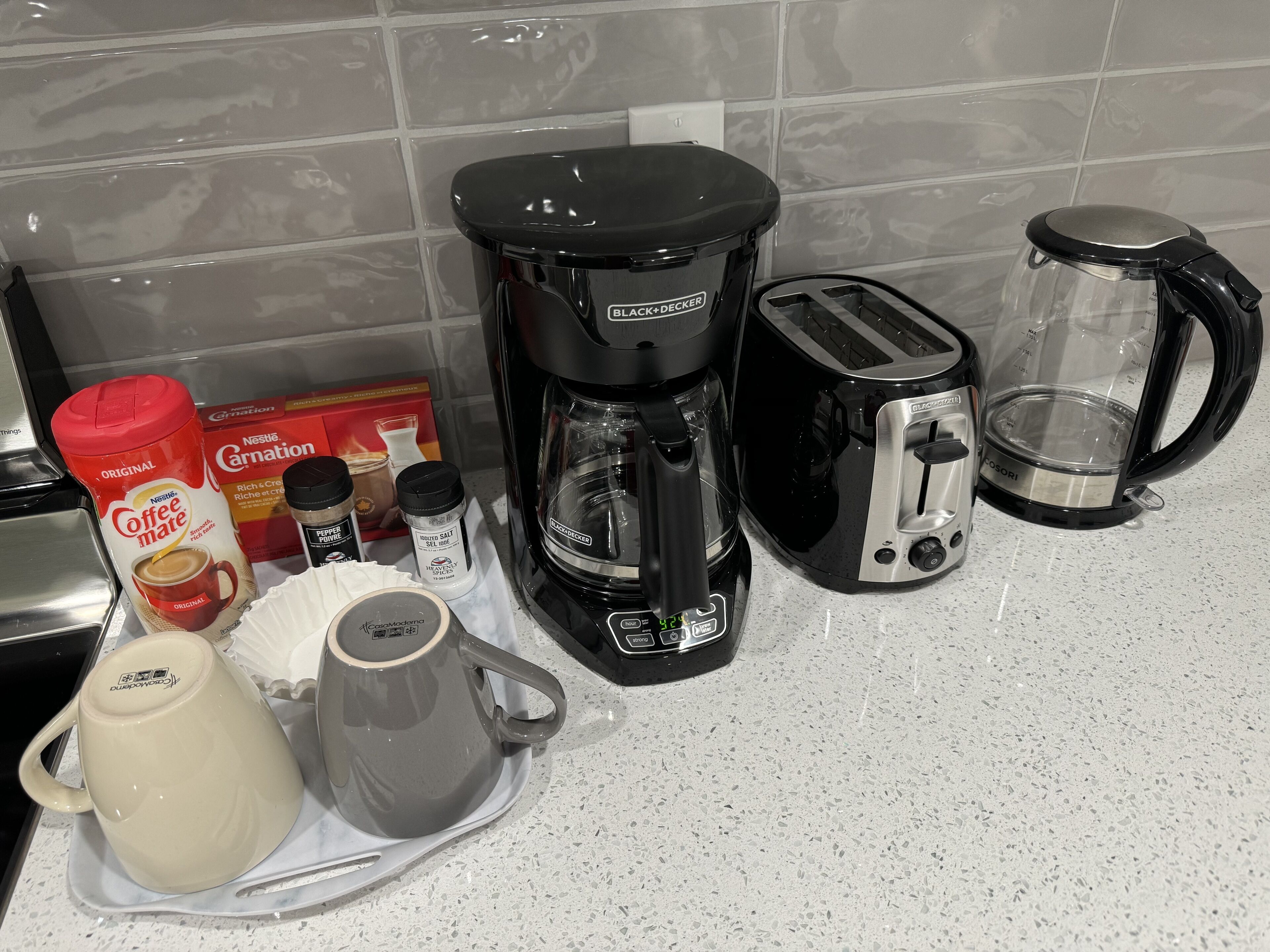 A kitchen counter with a coffee maker, toaster, kettle, and coffee mugs.
