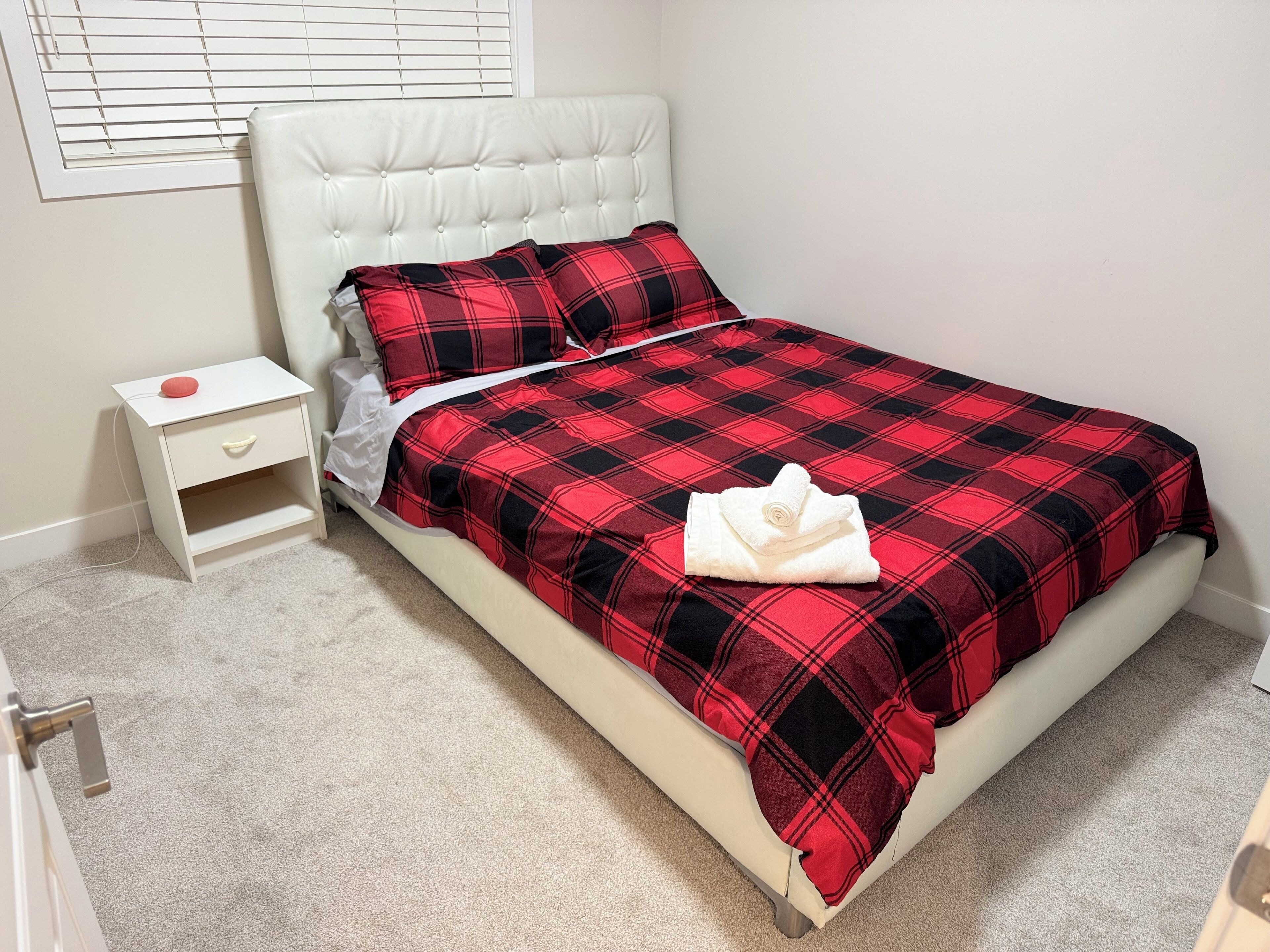 Una cama bien hecha con una colcha roja y negra a cuadros, una almohada blanca y una toalla blanca enrollada sobre la cama.
