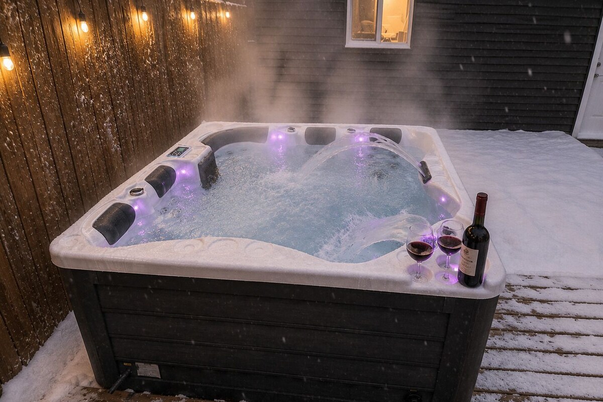 A hot tub with a control panel, surrounded by snow, and a bottle and glasses on the side.