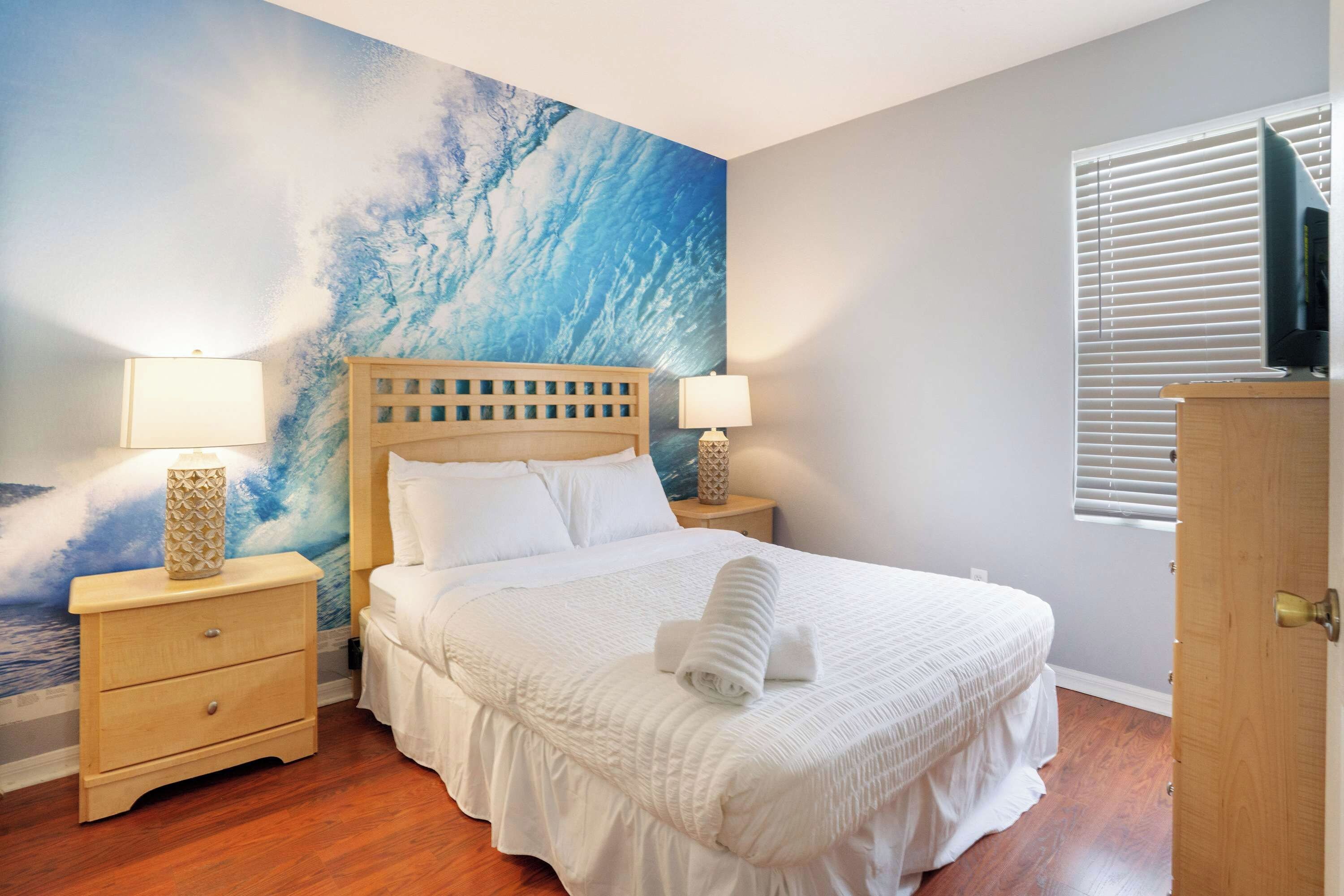 Een slaapkamer met een bed, twee nachtkastjes, een wanddecoratie, een raam met jaloezieën en een houten deur.