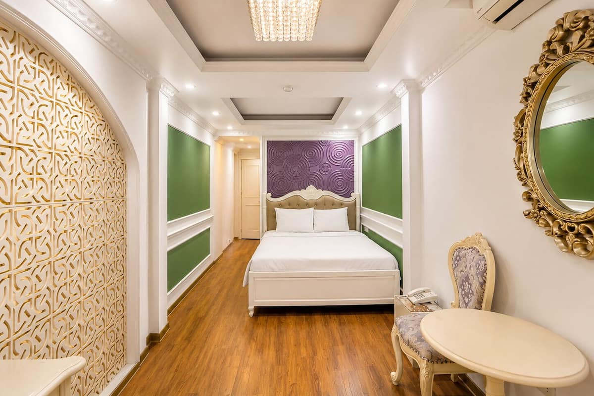 A hotel room with a bed, a chair, a small table, a decorative mirror, and a chandelier.
