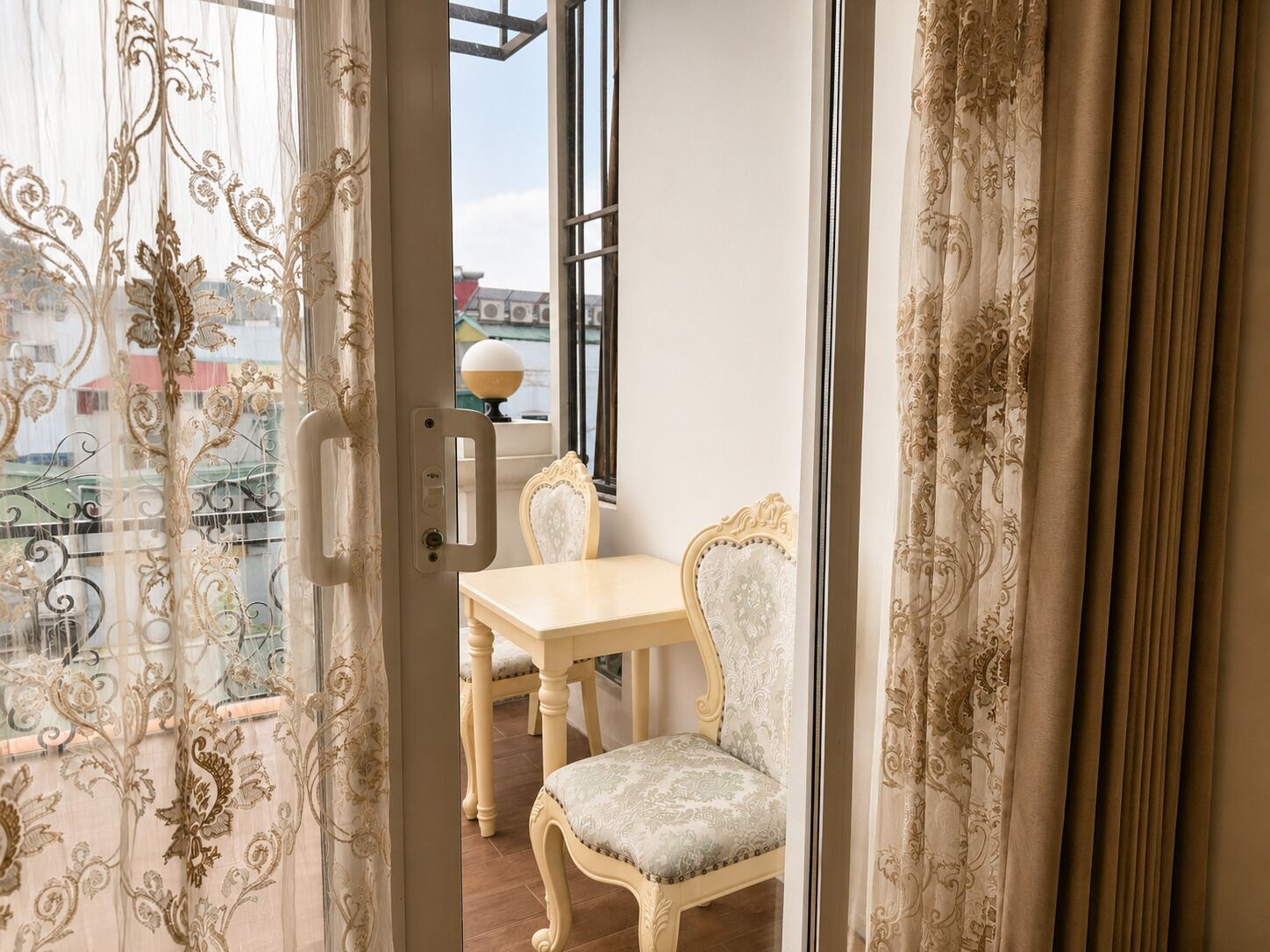 Un balcon avec une table et des chaises, une vue sur les immeubles et des rideaux décoratifs.