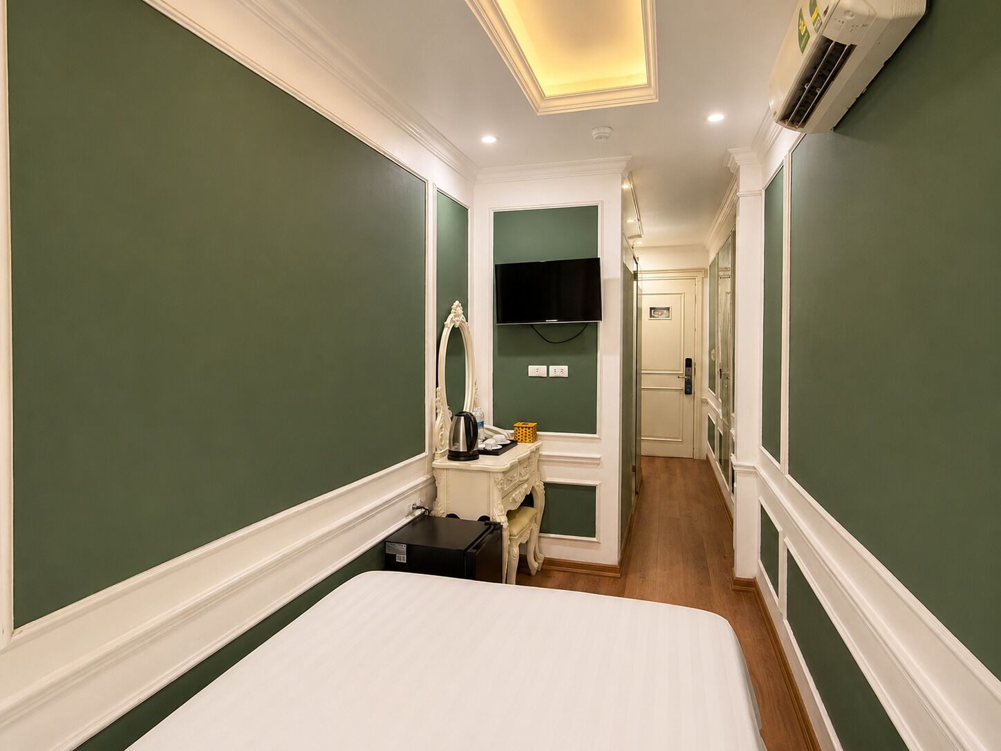 A hotel room with a bed, a desk with a mirror, a television, and an air conditioning unit.