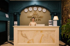 Un comptoir d’accueil avec des horloges indiquant l’heure de différentes villes, une enseigne indiquant « HANOI SILK CLASSY HOTEL & SPA », et une plaque nominative portant l’inscription « Lenovo ».