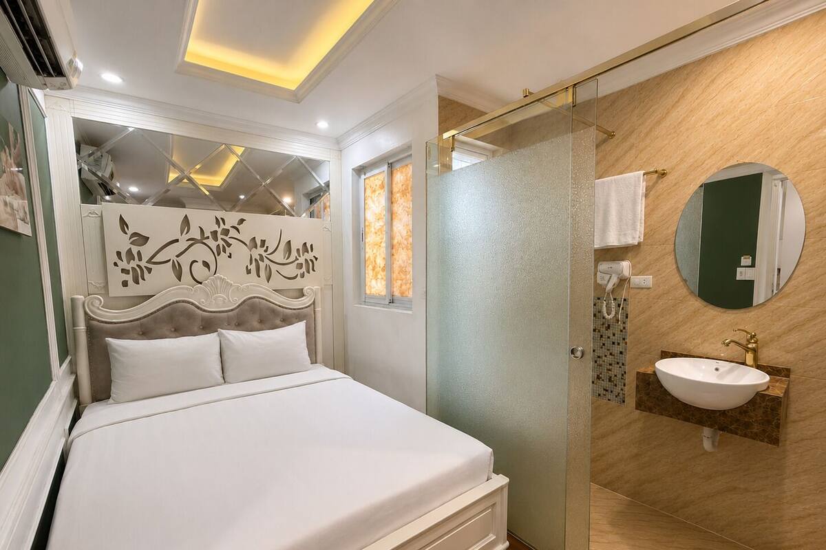 A bedroom with a bed, a glass shower enclosure, a bathroom with a sink and mirror, and a decorative wall panel.