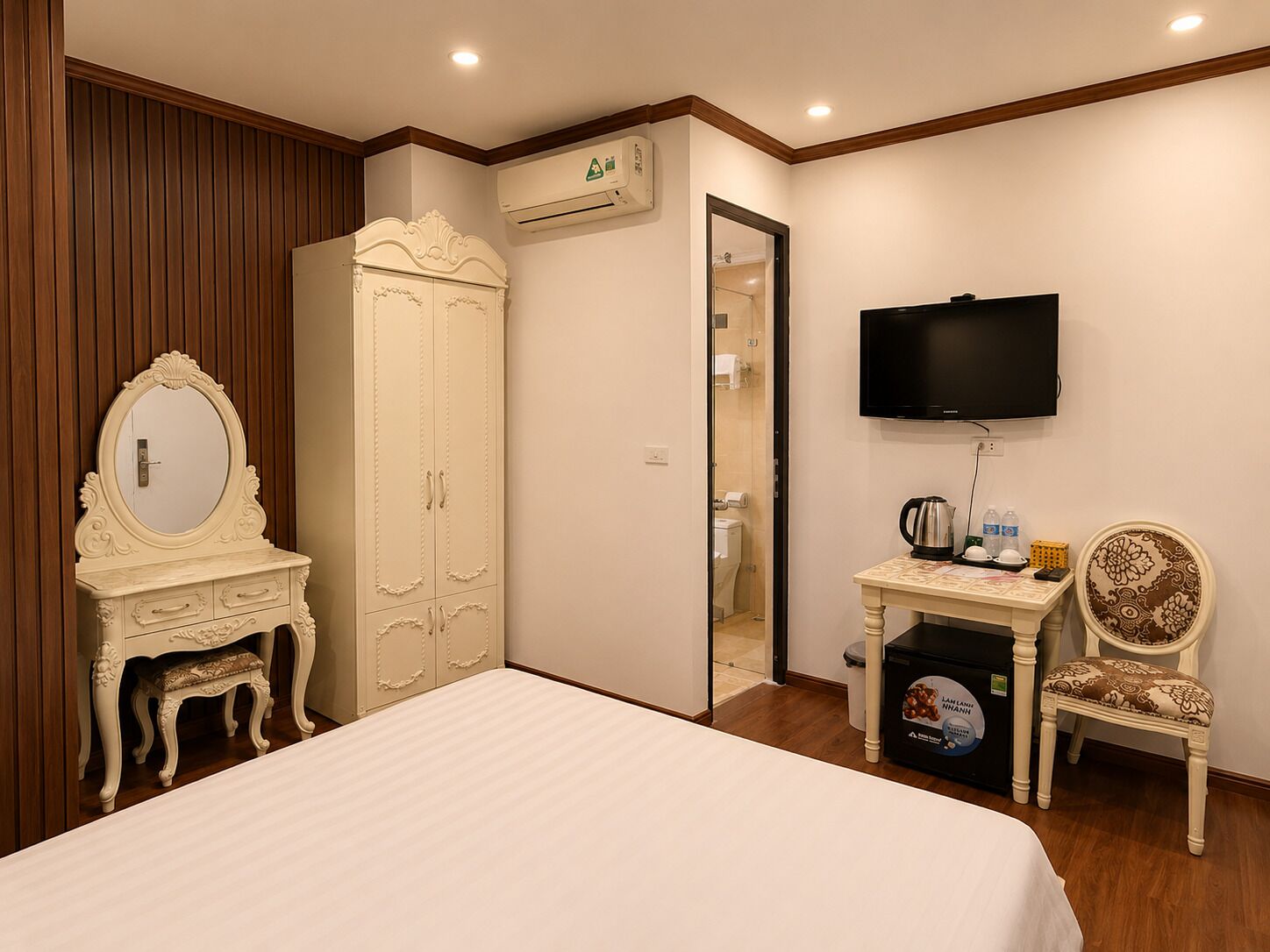 A bedroom with a bed, a vanity with a mirror, a wardrobe, a television, a small table with a kettle, and a chair.