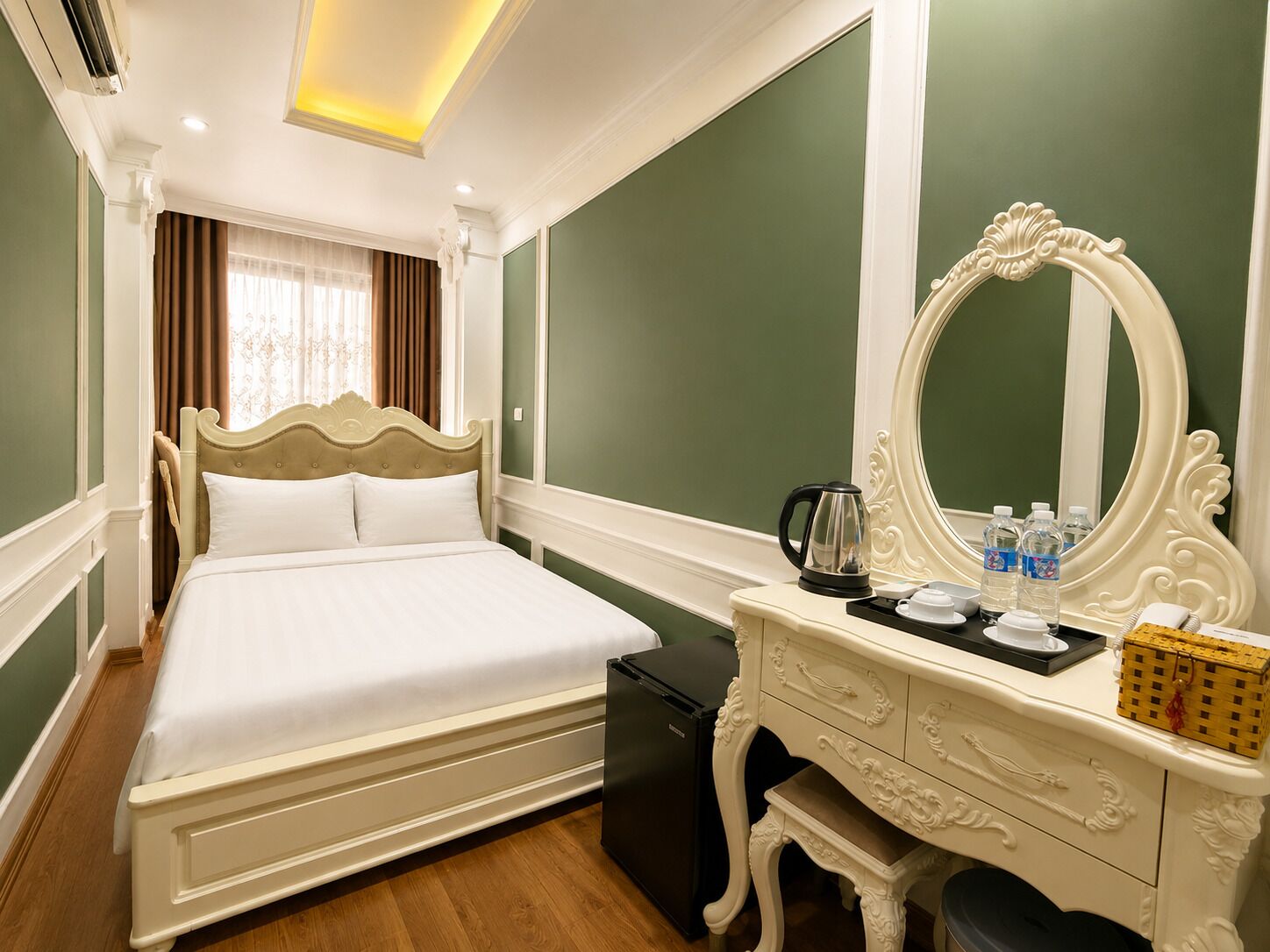 Quarto de hotel com uma cama grande, cômoda, espelho e pequena mesa com chaleira e xícaras.