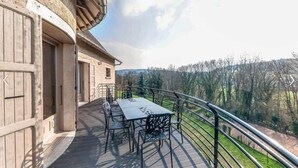 Una terraza de madera con una mesa y sillas, barandilla metálica y vistas a árboles y colinas.