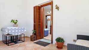 Una habitación con una cama, una mesa con mantel estampado, una planta en maceta, una puerta y un taburete pequeño.