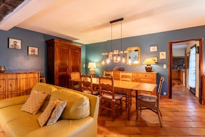 A dining area with a wooden table and chairs, a yellow sofa with patterned cushions, and a hanging light fixture with exposed bulbs.