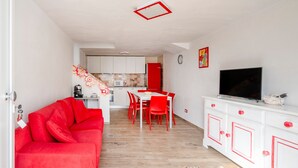 Um espaço de estar compacto com sofá vermelho, área de jantar, cozinha e televisão sobre um móvel branco.