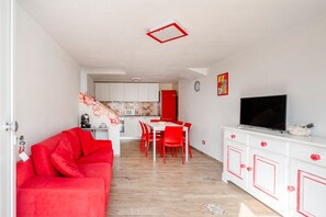 Un espacio habitable compacto con un sofá rojo, una zona de comedor, una cocina y un televisor sobre un mueble blanco.