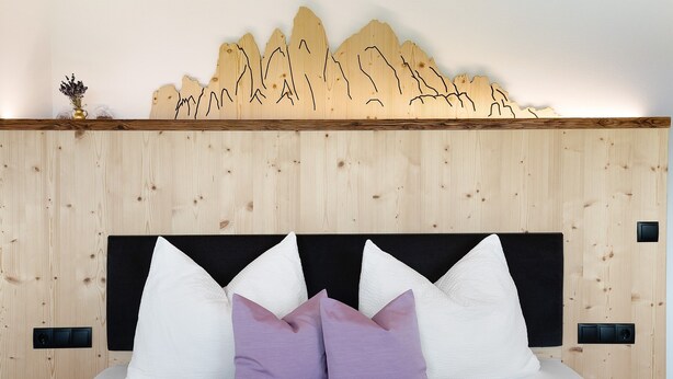 Cabecera de madera con diseño de montaña, cama con almohadas blancas y moradas, y un pequeño florero sobre la mesita de noche.