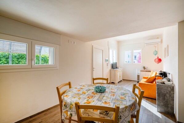 A dining area with a table and chairs, a kitchen area with a sofa, and a view of greenery outside.