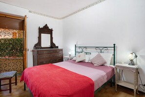 Camera da letto con un letto, una cassettiera, uno specchio, un comodino, una lampada e vista su piante attraverso una porta.