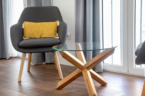 Une pièce moderne avec un fauteuil gris, une table basse en verre avec des pieds en bois, un plancher en bois et une fenêtre avec des rideaux.