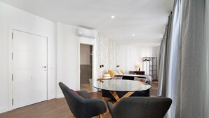 Un salon moderne avec une table à manger au plateau en verre, des chaises recouvertes d’un tissu gris, un canapé avec des coussins, une table d’appoint et un climatiseur fixé au mur.