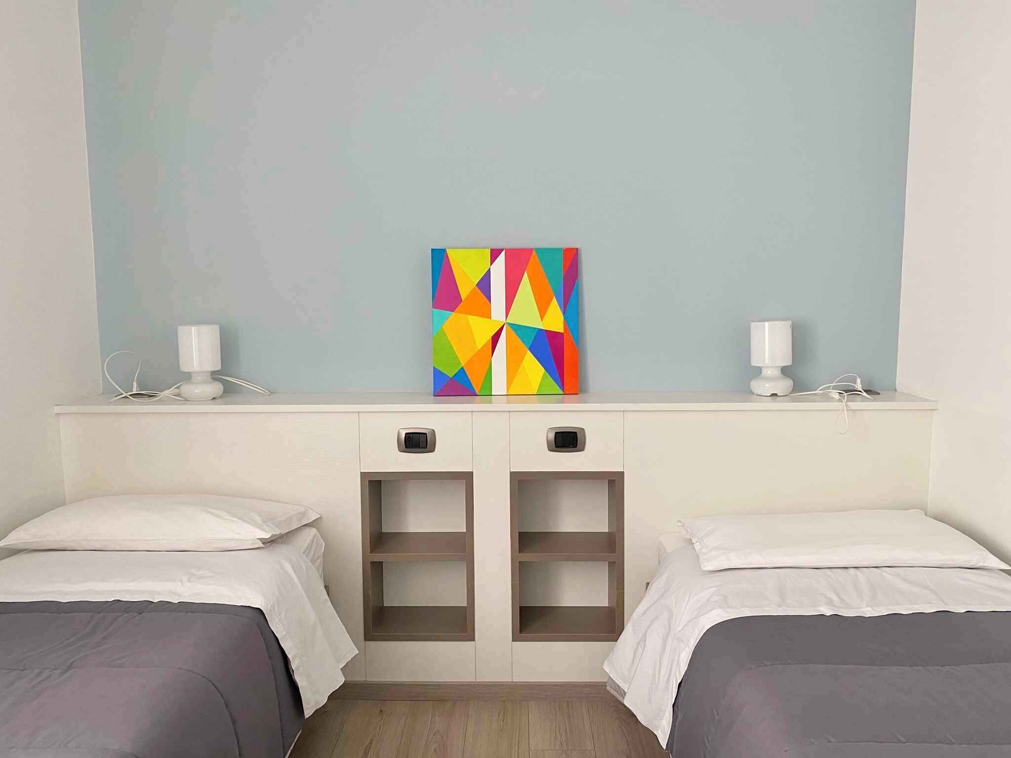 Een slaapkamer met twee bedden, een kleurrijk geometrisch schilderij en twee bedlampen.