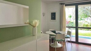 Ein Zimmer mit Esstisch, Stühlen, einer Pflanze in einer Vase, einem Deckenventilator und einem Fenster mit Blick auf Grünflächen.