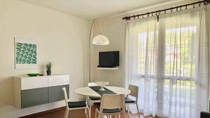 Una sala de estar moderna con comedor, televisor de pantalla plana en la pared y un ventanal con cortinas translúcidas.