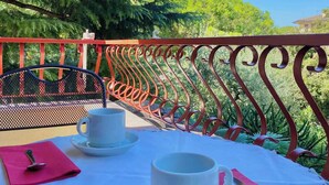 Ein Tisch ist gedeckt mit weißen Tassen und Untertassen, einer rosa Serviette und einem Löffel. Der Balkon hat eine rote Brüstung und bietet Blick auf Bäume und ein Haus.