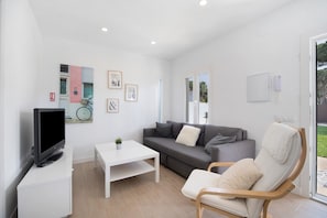 En moderne stue med et fladskærms-tv, et hvidt sofabord, en grå sofa, en hvid lænestol og en lille potteplante.