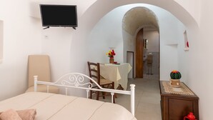 A bedroom with a bed, a TV mounted on the wall, a dining table with chairs, and an arched doorway leading to a bathroom. - Villa 'Trulletti Gioia' with Private Pool, Wi-Fi and Air Conditioning (Ceglie Messapica)