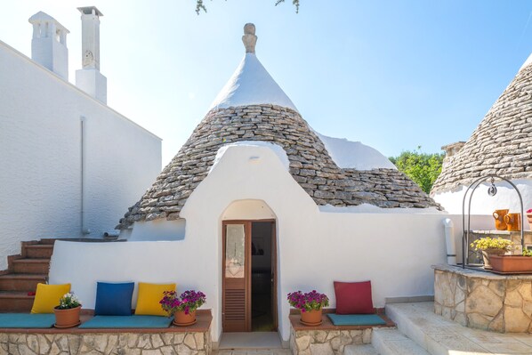 Un bâtiment blanc avec un toit en pierre, une porte et un petit porche agrémenté de coussins colorés et de plantes en pot.