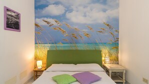A bed with a green headboard, two pillows, a nightstand with a lamp, and a wall painting of a beach scene.