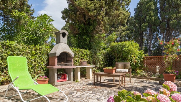 A patio with a stone-tiled floor, a green lounge chair, a wooden table, and a brick oven.