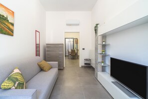 Ein modernes Wohnzimmer mit einem grauen Sofa, einem Flachbildfernseher und einem Essbereich im Hintergrund.